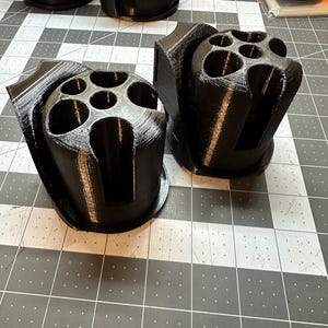 May include: Two black 3D-printed holders, each with a cylindrical design featuring multiple circular openings. The holders are designed to hold multiple items. The items are sitting on a gray and white grid surface.