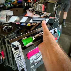 May include: A black and white film clapperboard with a black handle and white diagonal stripes. The clapperboard is open, revealing a green marker and a pink card. The clapperboard is used in the film industry to synchronize sound and picture.