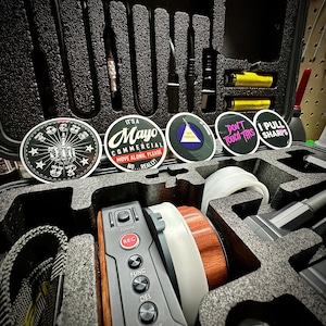 May include: A black case with foam inserts holds various equipment, including a camera control panel, lenses, and cables. Several round stickers with text are visible, including "F*** Up", "It's a Mayo Commercial", "Don't Touch This", and "I Pull Sharps".