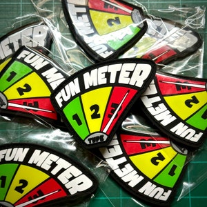 Puede incluir: Varios parches Fun Meter, empaquetados individualmente. Cada parche presenta un diseño de medidor con una aguja y el texto "FUN METER". El medidor está dividido en secciones verdes, amarillas y rojas, con los números 1, 2 y 3.