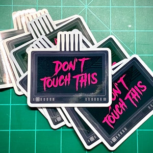 May include: A collection of stickers featuring a retro television design. The screen displays the text "DON'T TOUCH THIS" in pink, stylized lettering. The stickers are on a green cutting mat.