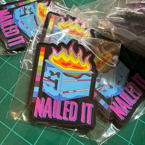 Könnte beinhalten: Mehrere schwarze Aufnäher in Plastikverpackung. Jeder Aufnäher zeigt eine blaue, orangefarbene und rosa Grafik eines brennenden Kuchens mit den Worten "NAILED IT" in Rosa. Die Aufnäher liegen auf einer grünen Schneidematte.