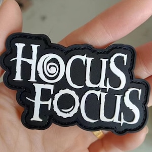Puede incluir: Un parche en blanco y negro con las palabras "Hocus Pocus" en una fuente estilizada. El parche tiene un borde negro y se sostiene en una mano.