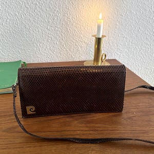 Könnte beinhalten: Eine rechteckige, dunkelbraune Umhängetasche mit Schlangenlederstruktur und einem dünnen, passenden Riemen. Die Tasche verfügt über ein goldfarbenes Metall-Logoemblem in der unteren linken Ecke, das ein stilisiertes P auf quadratischem Grund zeigt.