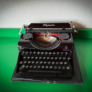 May include: A black Olympia Simplex typewriter with a glossy finish, set on a black leather case. The keys are white with black lettering. The word "Olympia" is printed on the top.