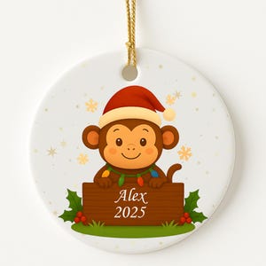 May include: A round Christmas ornament featuring a cartoon monkey wearing a Santa hat and holding a wooden sign that reads "Alex 2025". The monkey is decorated with Christmas lights and holly. The ornament has a gold string for hanging.