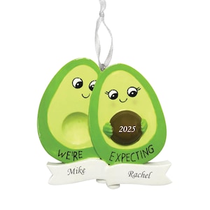 May include: A whimsical avocado-shaped ornament with a light green exterior and a brown pit. The ornament reads "We're Expecting" and "2025" on the pit. A banner below reads "Mike" and "Rachel". A white ribbon is attached for hanging.