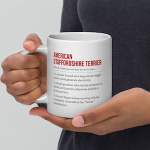 Può includere: Una tazza di ceramica bianca con la scritta "AMERICAN STAFFORDSHIRE TERRIER" in rosso e una definizione. La definizione include descrizioni umoristiche della razza. La tazza è tenuta da una persona.