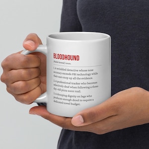 May include: White ceramic mug with the word "BLOODHOUND" in red, along with a definition. The definition includes three points about a detective, tracker, and drooling. The mug is held by a person.