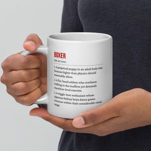 Könnte beinhalten: Weißer Keramikbecher mit dem Wort "BOXER" in Rot und einer Definition. Die Definition beschreibt einen Hund mit spielerischen Eigenschaften. Der Becher wird von einer Person gehalten.