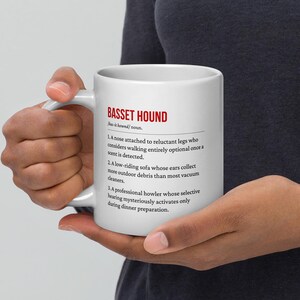May include: White ceramic mug featuring the text "BASSET HOUND" in red, with a humorous definition of the breed. The mug is held by a person, showcasing the text and design.