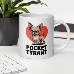 以下が含まれることがあります： 白いセラミックマグカップ。漫画のチワワのデザイン。チワワは赤いハートの背景で描かれています。「POCKET TYRANT」の文字が犬の下に黒で印刷されています。