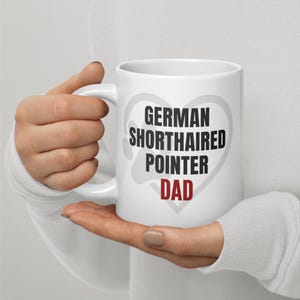 May include: White ceramic mug with the text "GERMAN SHORTHAIRED POINTER DAD" in black and red, with a gray heart-shaped design behind the text. The mug has a curved handle.