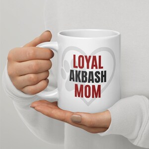 Puede incluir: Taza de cerámica blanca con las palabras "LOYAL AKBASH MOM" en letras rojas y negras, con un diseño de corazón y huellas de patas grises. La taza tiene un asa blanca.