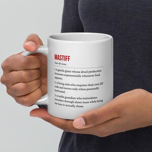 May include: White ceramic mug with the definition of "Mastiff" printed in black and red. The definition includes humorous descriptions of the dog breed. The mug is held by a person.