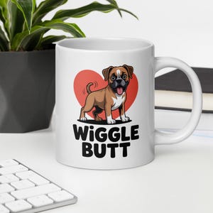 May include: White ceramic mug featuring a cartoon dog with brown and white fur, a red heart background, and the words "Wiggle Butt" in black. The mug has a curved handle and is on a white surface.