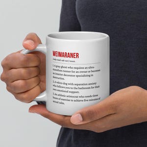 Op de afbeelding: Witte keramische mok met het woord "WEIMARANER" in het rood en een humoristische definitie. De mok bevat een definitie van een Weimaraner hond, inclusief zijn kenmerken. De mok wordt door een persoon vastgehouden.