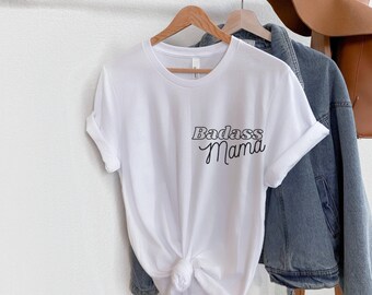 Puede incluir: Camiseta blanca con el texto "Badass Mama" en una fuente retro.