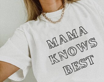Puede incluir: Camiseta blanquecina con el texto "MAMA KNOWS BEST" en letras negras desgastadas. Se ve un collar de cadena dorada. La camiseta es una prenda informal para el día a día.