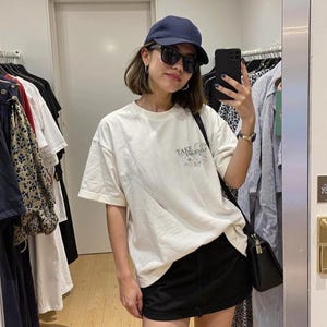 Puede incluir: Una persona con gorra azul marino, gafas de sol negras y una camiseta blanca con el texto "Take Care of Yourself". La persona tambi&eacute;n lleva una falda negra y un bolso negro.