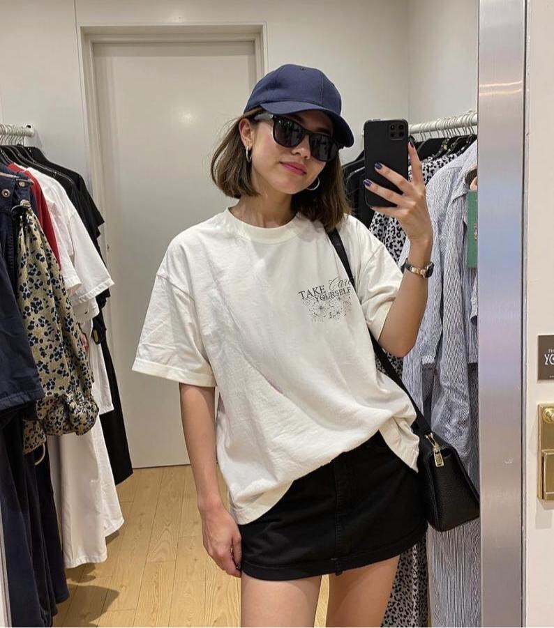 Puede incluir: Una persona con gorra azul marino, gafas de sol negras y una camiseta blanca con el texto "Take Care of Yourself". La persona tambi&eacute;n lleva una falda negra y un bolso negro.