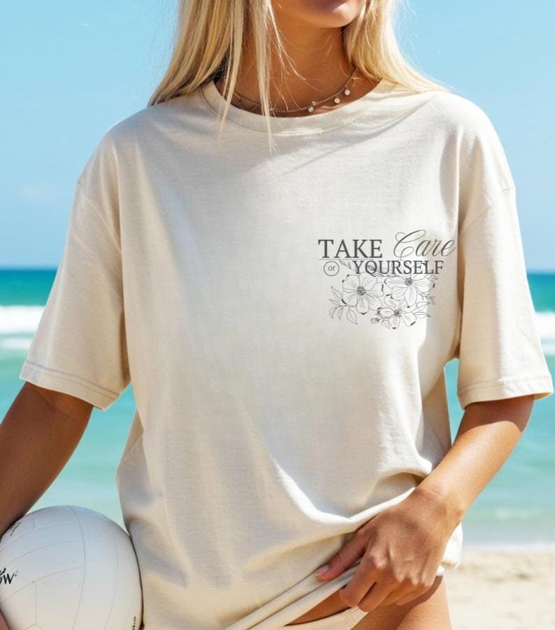 Puede incluir: Camiseta color crema con el texto "TAKE CARE OF YOURSELF" y un dise&ntilde;o floral. La camiseta es de corte holgado y tiene mangas cortas. Una persona sostiene un bal&oacute;n de voleibol blanco.