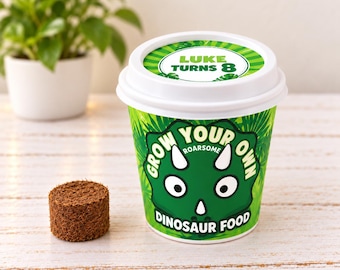 May include: A white plastic cup with a green dinosaur graphic and the text "GROW YOUR OWN ROARSOME DINOSAUR FOOD" on the side, and "LUKE TURNS 8" on the lid. A small, round, brown compressed soil disc sits next to the cup on a white surface.