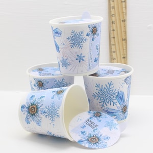 May include: A collection of white paper cups featuring blue snowflake and floral patterns. The cups are adorned with lids displaying matching designs and the text "AMELIE TURNS". The cups are decorated with blue crystals and the text "THANK YOU FOR COMING TO MY PARTY".