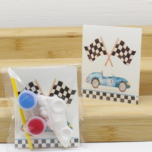May include: A craft kit for kids to paint a white race car. The kit includes two small paint pots, a paintbrush, and a yellow straw. The kit also includes a card with a blue race car and checkered flags.