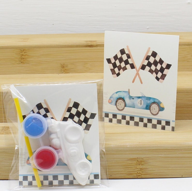May include: A craft kit for kids to paint a white race car. The kit includes two small paint pots, a paintbrush, and a yellow straw. The kit also includes a card with a blue race car and checkered flags.