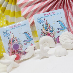 May include: Mermaid-themed party favor bags with thank you cards that say "Thank You for Coming to My Party". The bags contain white ceramic starfish, seashells, and a small paint set.