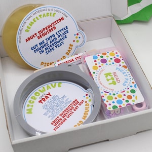 May include: A white box with a grey lid containing a craft kit for making a bead pick-up pad. The kit includes a clear plastic container with colourful beads, a pink plastic tray with a white label that says "Create Your Own Picky Pad Bead Kit", a white silicone tray with a label that says "Microwave Safe Tray", and a white label that says "Adult Supervision Required Cut me into little chunks and place in microwave safe tray".