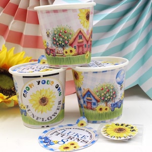 May include: Three small white cups with blue and white checked patterns and colourful images of sunflowers, houses, and tractors. The cups have labels that say "Grow Your Own Sunflower" and "Seeds". A blue and white circular sticker with the text "Luke Turns 8" is also visible.