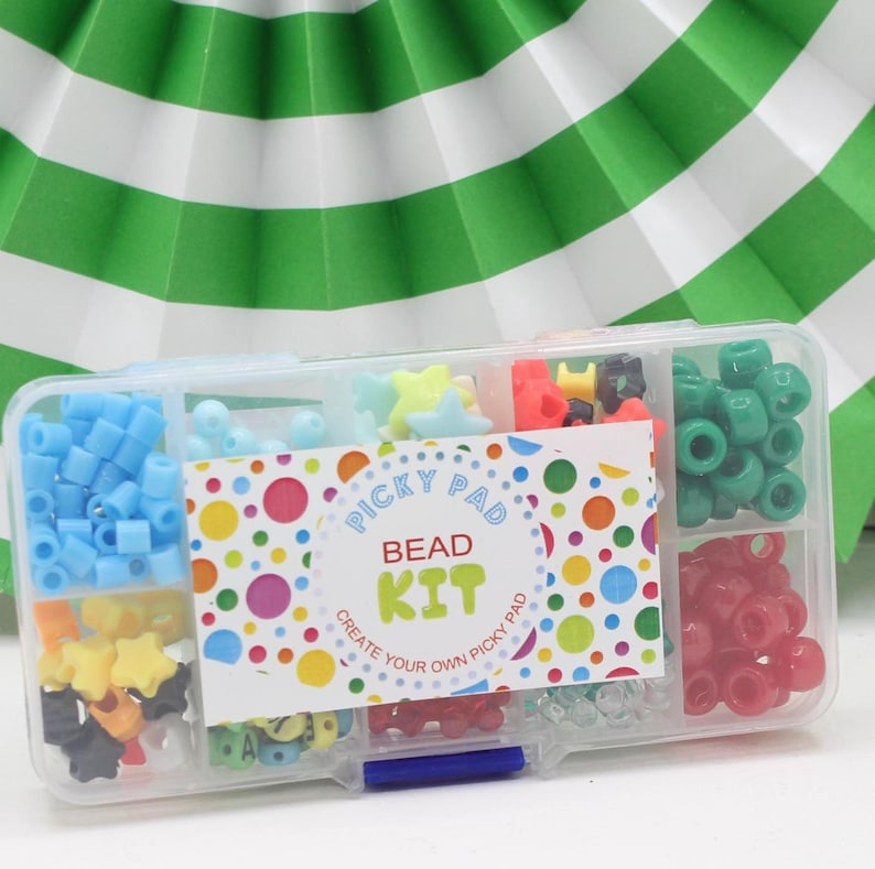 May include: A clear plastic container with a blue lid filled with colourful beads. The container has a label that says "Picky Pad Bead Kit Create Your Own Picky Pad".