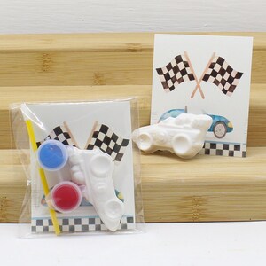 May include: A white ceramic race car with black checkered flags and two small paint pots, one red and one blue, with a yellow paintbrush.