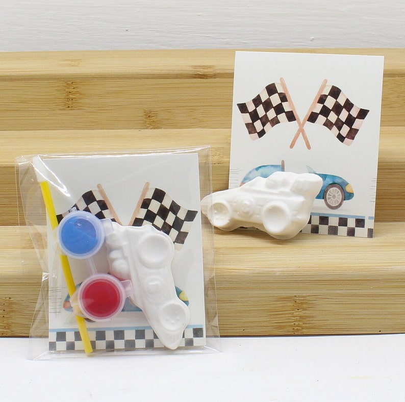 May include: A white ceramic race car with black checkered flags and two small paint pots, one red and one blue, with a yellow paintbrush.