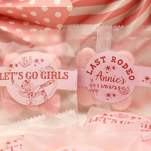 May include: Two cellophane bags filled with pink sweets, each sealed with a pink label. One label reads "Let's Go Girls" with a cowgirl illustration. The other says "Last Rodeo Annie's 11/06/25". The background is pink and white.