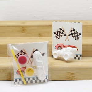 May include: A white ceramic race car figurine with red and yellow paint pots and a paintbrush in a clear plastic bag. The bag also includes a card with a red race car and two black and white checkered flags.