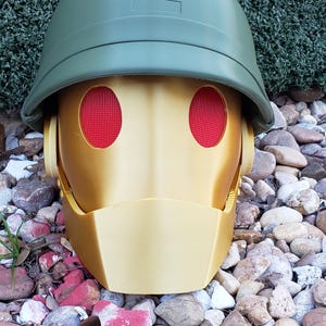 May include: A gold robot head with red eyes, wearing a green helmet with the number "2" on it. The robot head is sitting on a bed of small rocks.