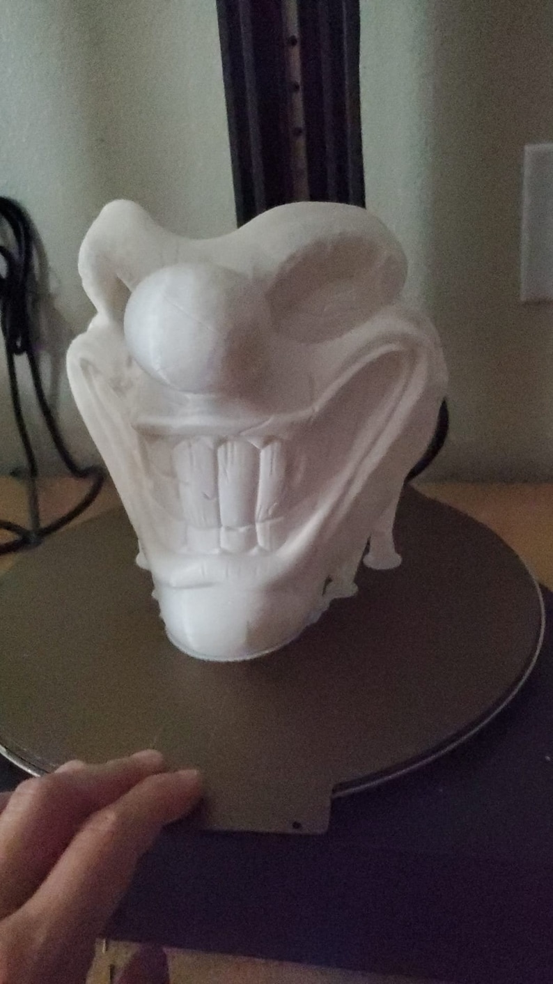 May include: A white 3D printed model of a grinning face with large teeth. The model is sitting on a black circular base.