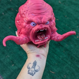 May include: A pink, grotesque, and angry-looking Krang head figurine with purple accents. The open mouth reveals teeth and a red interior. The figurine is held up against a green background.