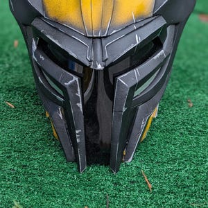 May include: A black and yellow helmet with a distinctive design, featuring angular shapes and a textured finish. The helmet has a weathered appearance with visible wear and tear. The helmet is resting on a green surface.