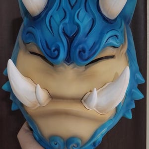 May include: A blue and tan Oni mask with white horns and tusks. The mask features intricate swirling details and closed eyes. The mask is held up by a hand.