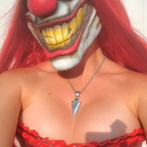 May include: A woman wearing a red and white clown mask with a large red nose and a wide, toothy grin. She is also wearing a red corset with black lace trim and a silver necklace with a pointed pendant.