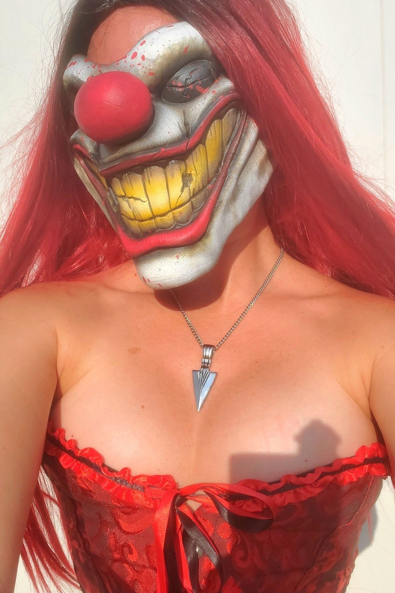 May include: A woman wearing a red and white clown mask with a large red nose and a wide, toothy grin. She is also wearing a red corset with black lace trim and a silver necklace with a pointed pendant.
