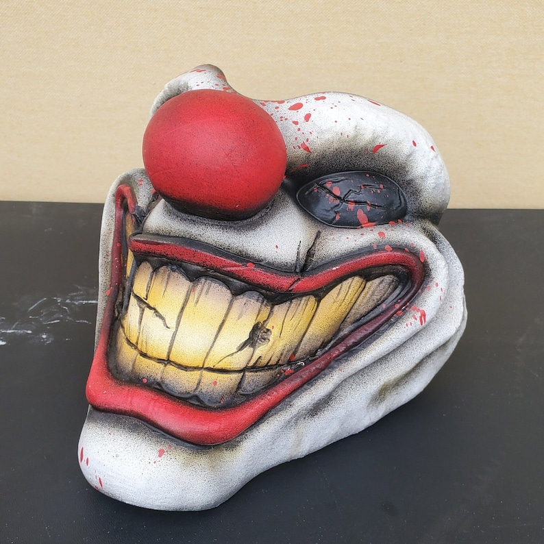 May include: A white ceramic clown mask with a red nose and a wide, toothy grin. The mask has black paint splatters and a single black eye.