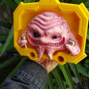 Könnte beinhalten: Eine pinkfarbene und gelbe 3D-gedruckte Figur eines Monsters mit einem zahnigen Grinsen. Das Monster hält in jeder Hand ein kleines, goldfarbenes Objekt.