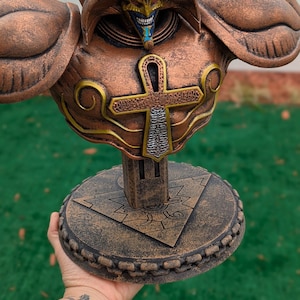 Puede incluir: Estatua detallada de color bronce de un personaje con casco y armadura estilizados. La estatua presenta detalles intrincados, incluyendo un emblema en forma de cruz y una base circular con patrones geométricos. La estatua se sostiene en una mano.