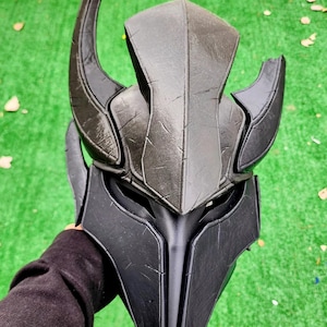 Puede incluir: Un casco negro y gris con una superficie texturizada y un diseño con cuernos. El casco tiene una barbilla puntiaguda y se sostiene sobre un fondo verde. El casco parece estar hecho de plástico o un material similar.