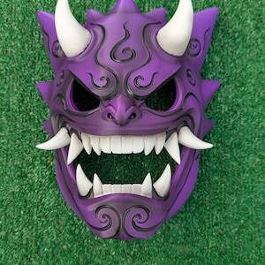 May include: A purple Oni mask with white horns and fangs. The mask features intricate swirling details and sharp, angular features. The mask is designed with a fierce expression, perfect for cosplay or display.
